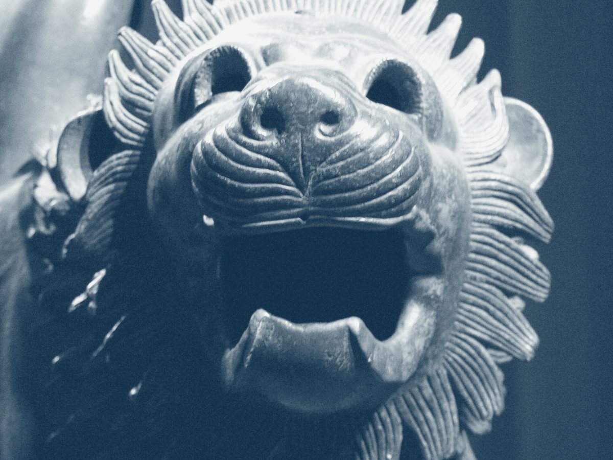 A close photograph of the bronze lion's head, mouth open, mane in flame-shaped locks.